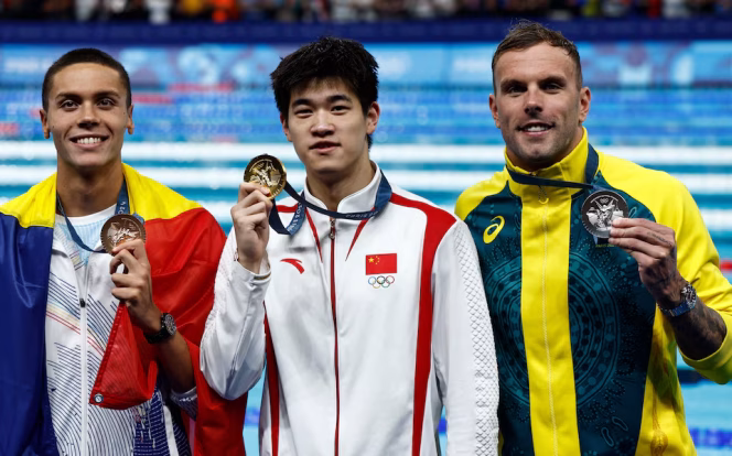 
					Paris 2024 Olympics - Swimming - Men's 100m Freestyle Victory Ceremony - Paris La Defense Arena, Nanterre, France - July 31, 2024. Gold medallist Zhanle Pan of China celebrates on the podium with silver medallist Kyle Chalmers of Australia and bronze medallist David Popovici of Romania. REUTERS/Clodagh Kilcoyne 